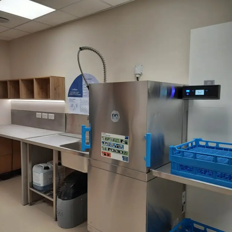 Dans un espace cuisine impeccable, un lave-vaisselle professionnel en acier inoxydable doté d’un panneau de commande est installé à côté d’un plan de travail muni d’étagères et d’un égouttoir à vaisselle bleu, illustrant l’avancement des travaux à la Maison de l’Éducation thérapeutique et reflétant son engagement en faveur de l’éducation thérapeutique dans un espace moderne et fonctionnel.