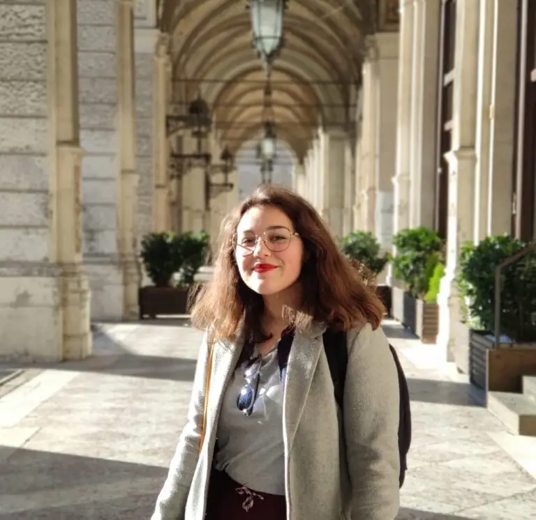 Une femme portant des lunettes et un sac à dos se tient debout, souriante, sous une passerelle voûtée richement décorée, aux colonnes de pierre et bordée de plantes, peut-être en route pour présenter son projet professionnel en chirurgie dentaire à la Bourse des donateurs.