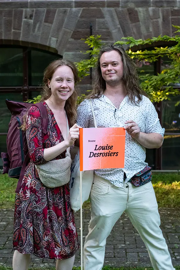 Deux personnes se tiennent à l'extérieur, sur un chemin pavé, souriantes, tenant une pancarte orange sur laquelle on peut lire « Louise Desrosiers ». À l'arrière-plan, on aperçoit un bâtiment en briques et de la verdure, pour célébrer leur sélection en tant que lauréats 2023 de la bourse Louise Desrosiers.