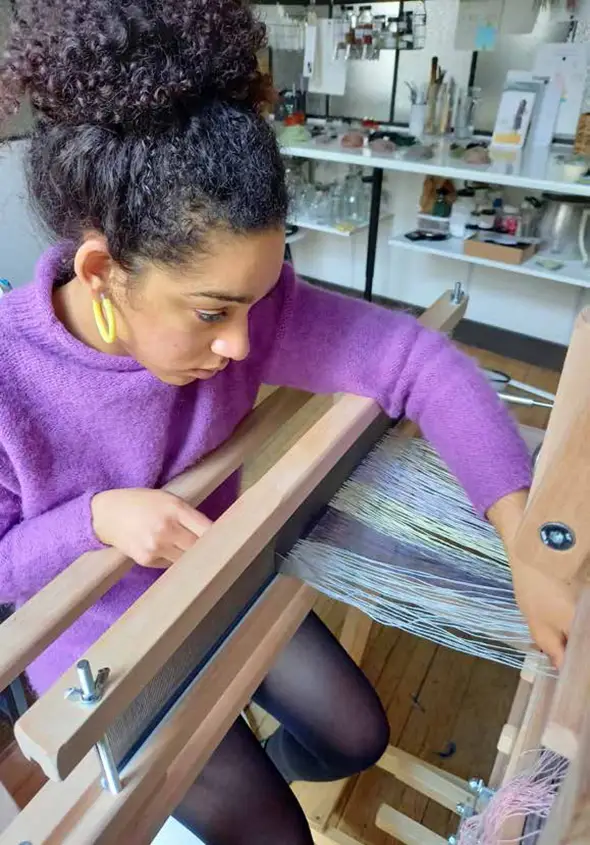 Une femme vêtue d'un pull violet, peut-être l'une des diplômées de l'HEAR, manie un métier à tisser dans un atelier bien éclairé, avec des étagères et du matériel en arrière-plan, mettant en valeur les compétences qu'elle a acquises grâce à divers projets.