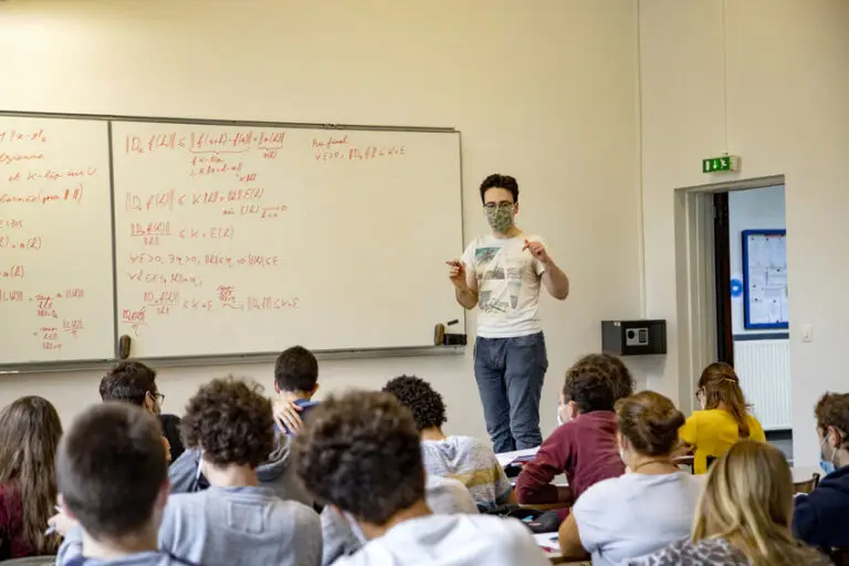En 2020, un enseignant portant un masque se tient devant sa classe et explique un cours de mathématiques au tableau blanc à des élèves assis à leurs pupitres, dont certains prennent des notes ; bon nombre d’entre eux peuvent fréquenter cet établissement grâce aux bourses accordées par de généreux donateurs.