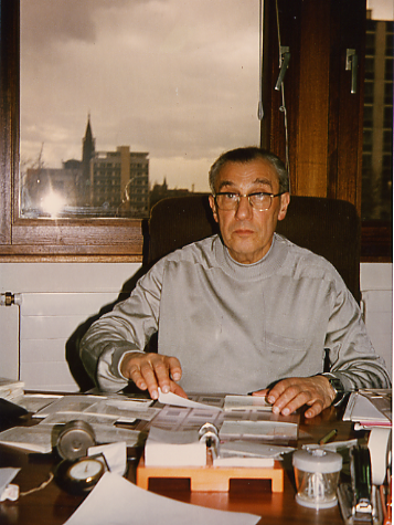 Un homme d’un certain âge, portant des lunettes, est assis à un bureau sur lequel sont disposés des papiers et des fournitures de bureau ; il regarde vers la caméra. Derrière lui, à travers la fenêtre, on aperçoit un paysage urbain qui rappelle Strasbourg, évoquant l’atmosphère universitaire où des personnalités telles que Paul Mandel ont apporté des contributions majeures à la neurochimie.