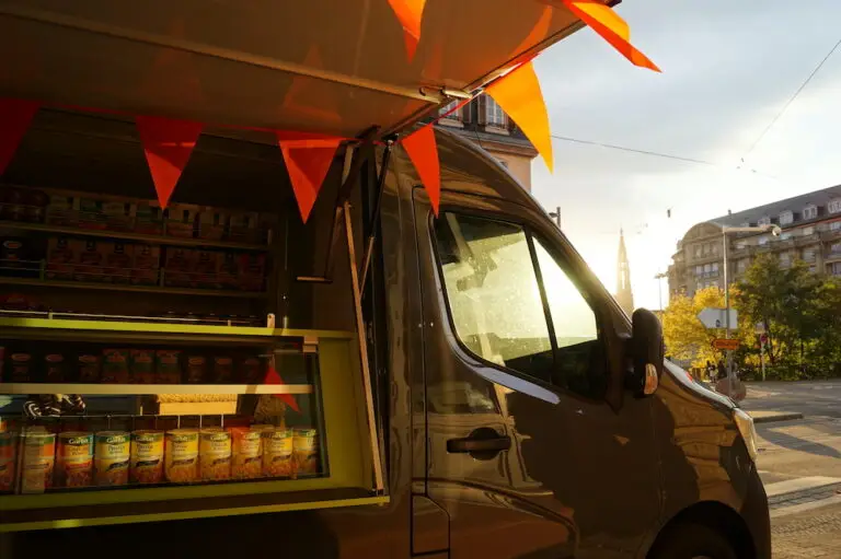 Un food truck, sur lequel sont exposées des boîtes de conserve et qui fait partie de l'initiative Agora'Truck, est garé dans une rue de la ville au coucher du soleil ; des drapeaux orange sont suspendus au-dessus de la vitrine, symbolisant sa vocation d'épicerie solidaire.