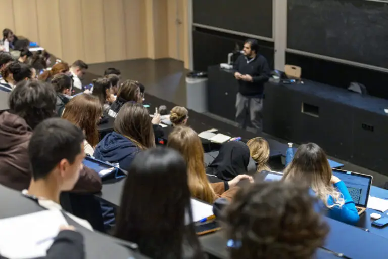 Les étudiants sont assis dans un amphithéâtre et écoutent un professeur qui s'exprime à l'avant de la salle, près d'un tableau noir, leurs cahiers et ordinateurs portables posés sur leurs pupitres, tandis qu'ils abordent des sujets tels que les assurances-vie, les donations et les legs dans le cadre de la planification successorale.