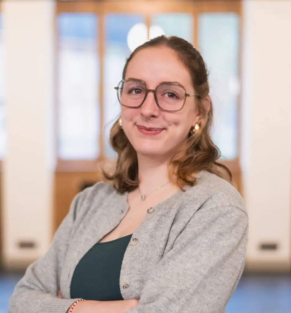 Une femme à lunettes aux cheveux châtain clair, vêtue d'un gilet gris, pose les bras croisés dans un intérieur bien éclairé à « La Fondation », avec des fenêtres floues en arrière-plan.