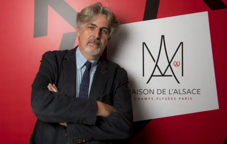 Un homme aux cheveux gris et à la barbe, les bras croisés, se tient debout devant l'enseigne « Maison de l'Alsace » sur fond rouge, incarnant la résilience face à la crise sanitaire qui a touché aussi bien les étudiants que les communautés.