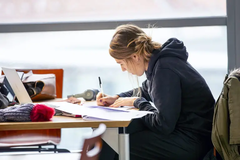 Une personne est assise à une table, les jambes croisées, en train d'écrire dans un cahier ; à côté d'elle se trouvent un ordinateur portable, des documents relatifs à des dons et des effets personnels.