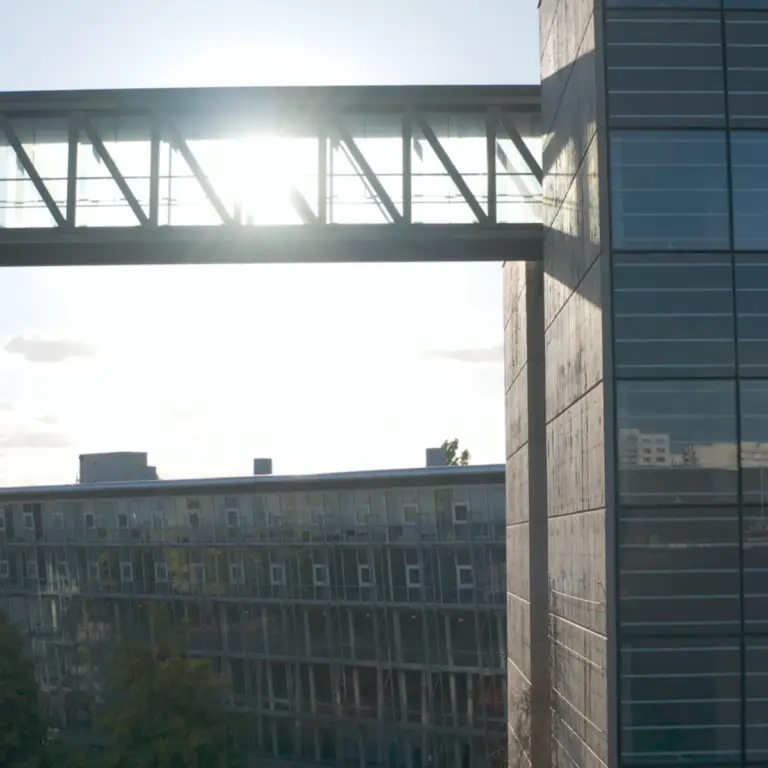 Une passerelle vitrée relie deux bâtiments modernes ; la lumière du soleil pénètre par les fenêtres du haut, tandis qu’un autre bâtiment aux façades vitrées se profile à l’arrière-plan — témoignage de l’engagement de Régis Bello envers son personnel et de son dévouement au mécénat pour favoriser l’innovation architecturale contemporaine.