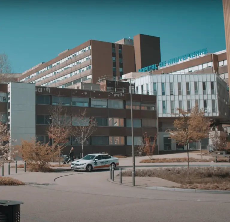 Un complexe hospitalier composé de plusieurs bâtiments modernes, avec l’enseigne « Hospital de Vall d’Hebron » bien visible et un taxi garé devant, entouré d’arbres sous un ciel dégagé : un pôle dédié à la santé, où des initiatives telles que le mécénat contribuent à soutenir des soins médicaux de pointe.