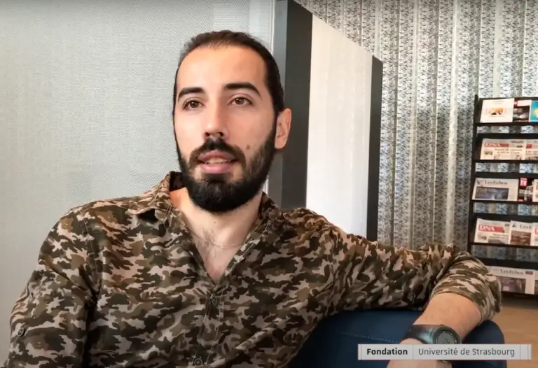 Un homme barbu, portant une moustache et une chemise de camouflage, est assis sur une chaise bleue dans une pièce moderne aux murs à motifs, avec des porte-revues en arrière-plan. Cet étudiant, Bachar Damour, est un étudiant réfugié à l'Université de Strasbourg.