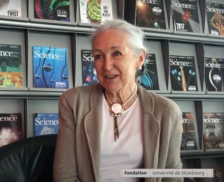 Une femme d’un certain âge est assise devant une étagère remplie de numéros du magazine *Science*. Elle porte un gilet beige et une chemise blanche, et ses cheveux gris sont tirés en arrière. Une étiquette indique « Fondation Université de Strasbourg ». Anne Becker, mécène engagée dans la recherche sur les maladies neurodégénératives, incarne l’engagement et la générosité à travers son soutien à cette fondation.