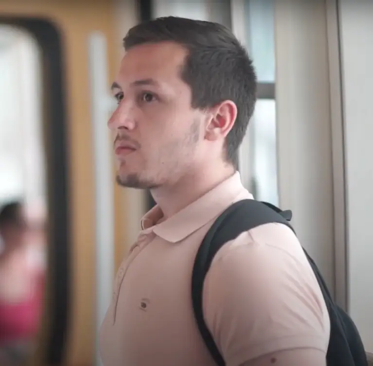 Un jeune homme aux cheveux bruns courts et à la barbe soigneusement taillée, bénéficiaire de la bourse des donateurs, porte un polo rose pâle et un sac à dos noir ; debout dans un train, il s'apprête à partager son témoignage.