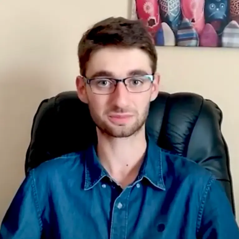 Un jeune homme à lunettes, portant une barbe soigneusement taillée, est assis sur une chaise noire, vêtu d'une chemise bleue boutonnée. Une œuvre d'art colorée est accrochée au mur beige derrière lui, tandis qu'il partage son témoignage sur sa vie d'étudiant boursier à la recherche d'opportunités au-delà des frontières.