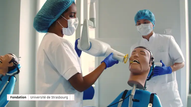 Deux personnes en tenue médicale s'entraînent à réaliser des actes dentaires sur un mannequin à l'aide de matériel dentaire, dans un cadre de formation clinique à la Faculté de chirurgie dentaire de l'Université de Strasbourg. Le texte figurant sur l'image indique : « Fondation Université de Strasbourg ».