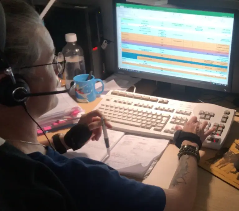 Une personne équipée d'un casque audio utilise un ordinateur tout en consultant un cahier de notes ; un grand écran affiche une feuille de calcul. On aperçoit divers objets de bureau, notamment une tasse bleue, une bouteille d'eau et des stylos : une scène typique des Hôpitaux universitaires de Strasbourg pendant la rénovation de leur standard téléphonique.