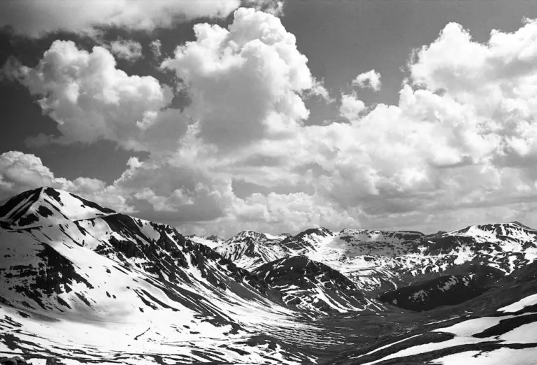Photo en noir et blanc représentant des montagnes enneigées sous un ciel parsemé de gros nuages cotonneux, qui rend compte de la grandeur sereine de la nature avec la même passion pour les sciences de la vie qui anime l'exploration et la découverte.