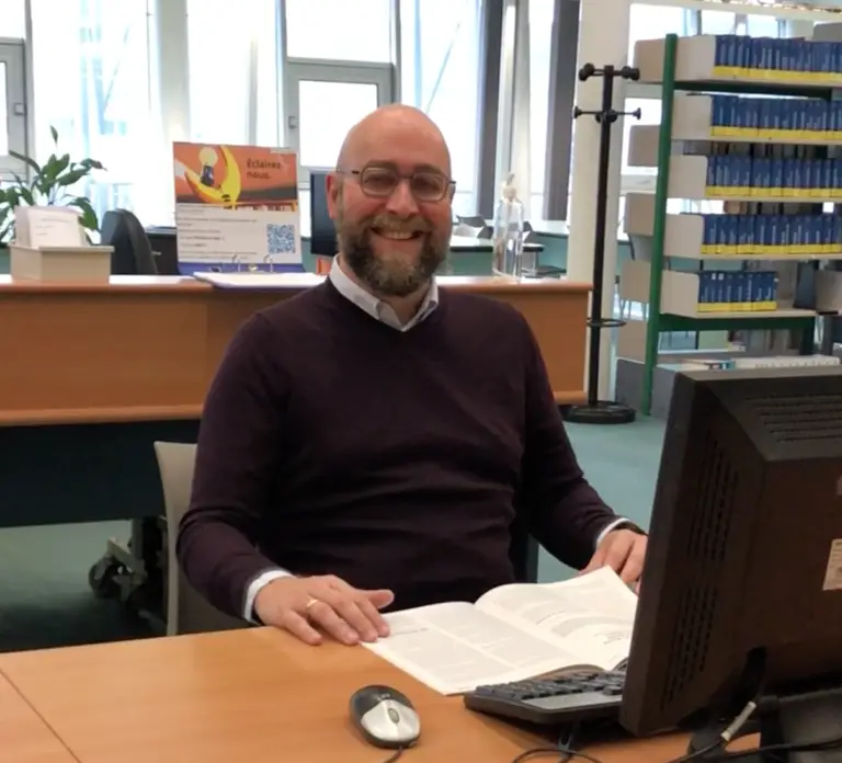 Un homme barbu et portant des lunettes est assis à un bureau, devant un livre ouvert et un ordinateur, dans une bibliothèque ou un bureau ; il sourit à la caméra, peut-être en train de se préparer à une soutenance de thèse ou de relire les travaux de Pascal Simon Doutreluingne.