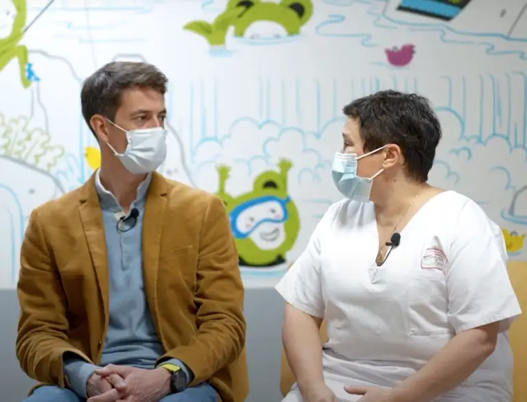 Un homme vêtu d'une veste marron et une femme en tenue médicale sont assis face à face, tous deux portant un masque, avec en arrière-plan une fresque murale colorée sur le thème de l'enfance — une scène qui illustre les projets collaboratifs soutenus par le mécénat au sein des Hôpitaux universitaires de Strasbourg.