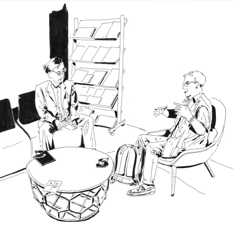 Deux personnes sont assises face à face dans un coin salon et discutent, engagées dans une conversation approfondie sur l'enseignement supérieur et la recherche, avec entre elles une bibliothèque et une table ronde sur laquelle sont posées des tasses et des feuilles.