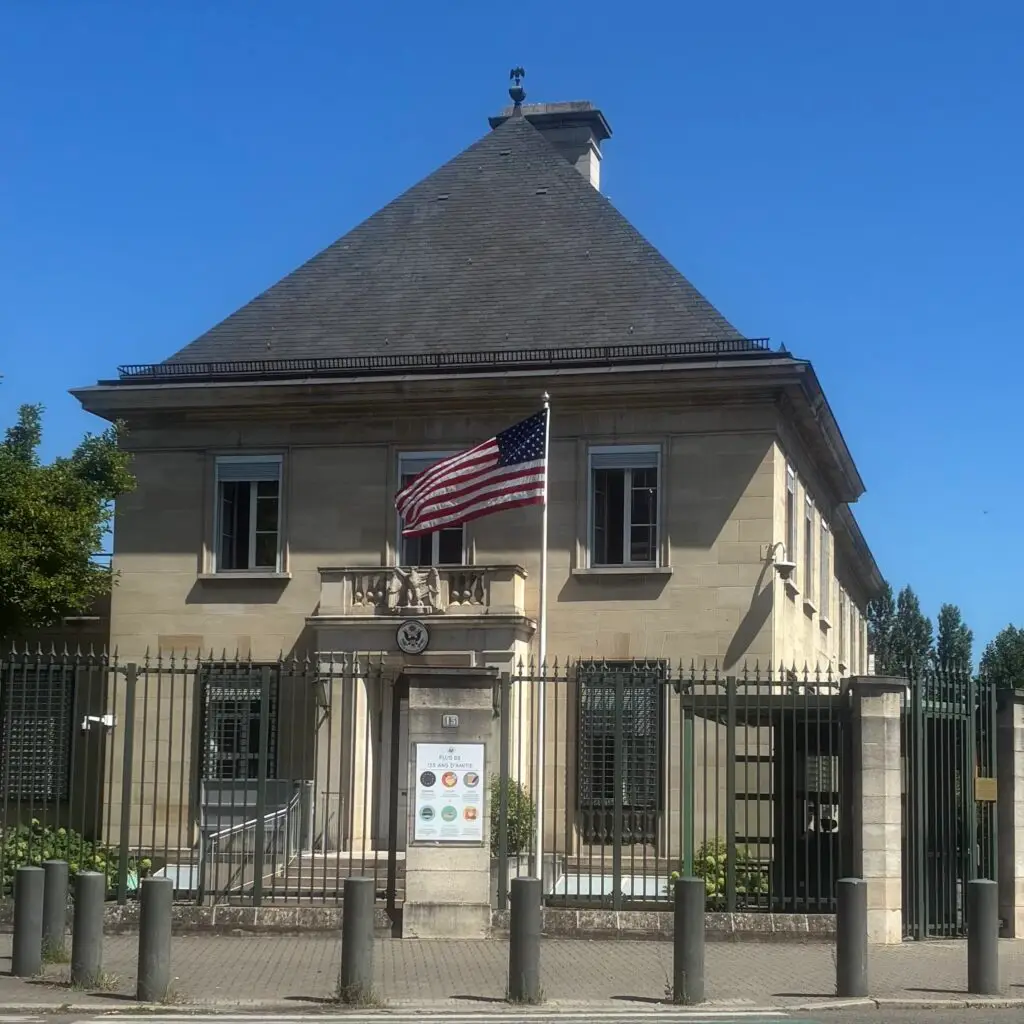 Un bâtiment en pierre beige au toit triangulaire, entouré d'une clôture, devant lequel flotte un drapeau américain. Sous un ciel bleu, près de l'entrée, un panneau orné d'emblèmes accueille les visiteurs souhaitant faire un don ou donner depuis l'étranger.