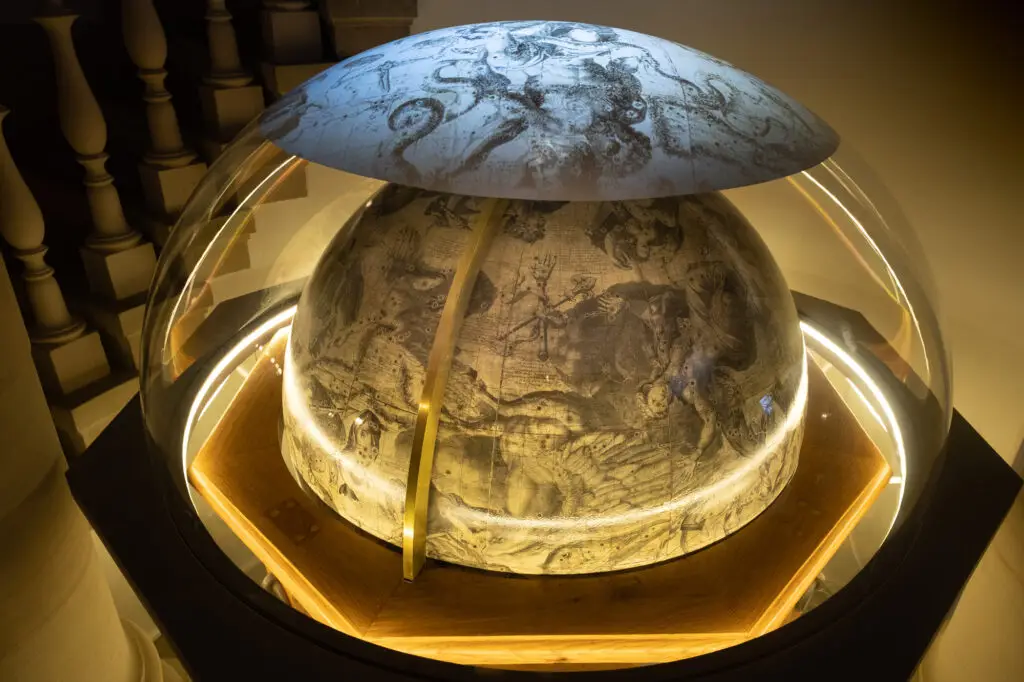 Un globe céleste en forme de dôme, sur la surface duquel sont gravées les constellations, est exposé sous une vitrine et éclairé par des lumières tamisées dans un cadre muséal, invitant les visiteurs à réfléchir aux nombreuses « formes de dons » — dons de savoir, d’émerveillement et de découverte — que représentent de tels objets scientifiques.