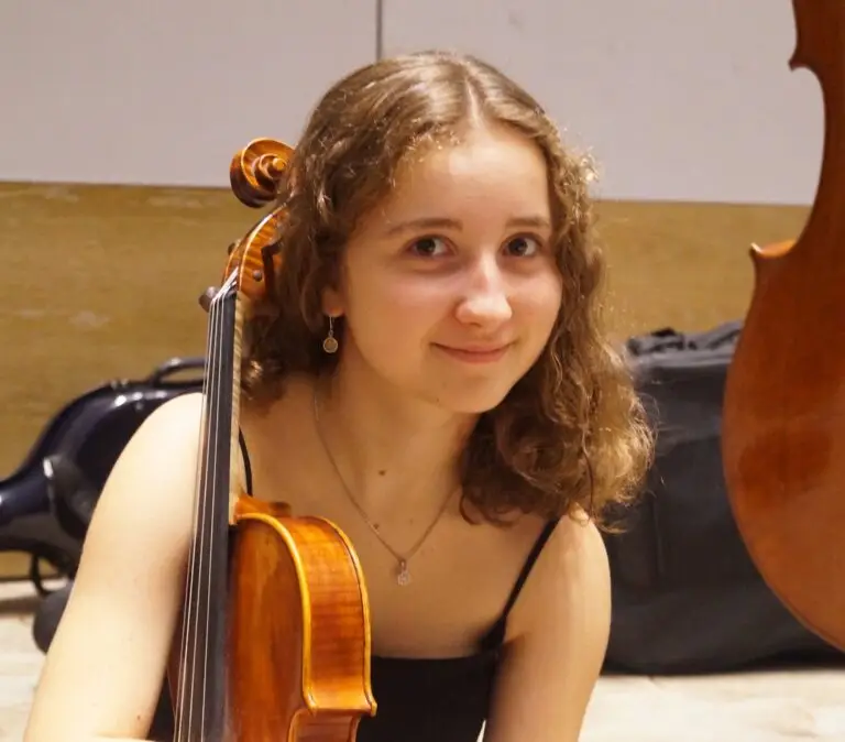 Dans une salle de répétition, une jeune femme aux cheveux bouclés tient un violon et sourit à la caméra, prête à partager son témoignage sur l'obtention de la bourse des donateurs. Elle s'appelle Aimée-Luce Ponza.