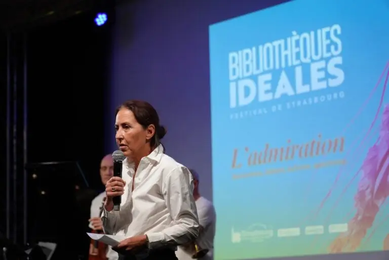 Sur la scène du Festival des Bibliothèques Idéales de Strasbourg, avec en arrière-plan un écran bleu dédié à l'événement, une femme vêtue d'une chemise blanche s'exprime au micro. Elle présente son livre « Homo Medicus » et partage ses réflexions avec le personnel soignant présent dans le public.