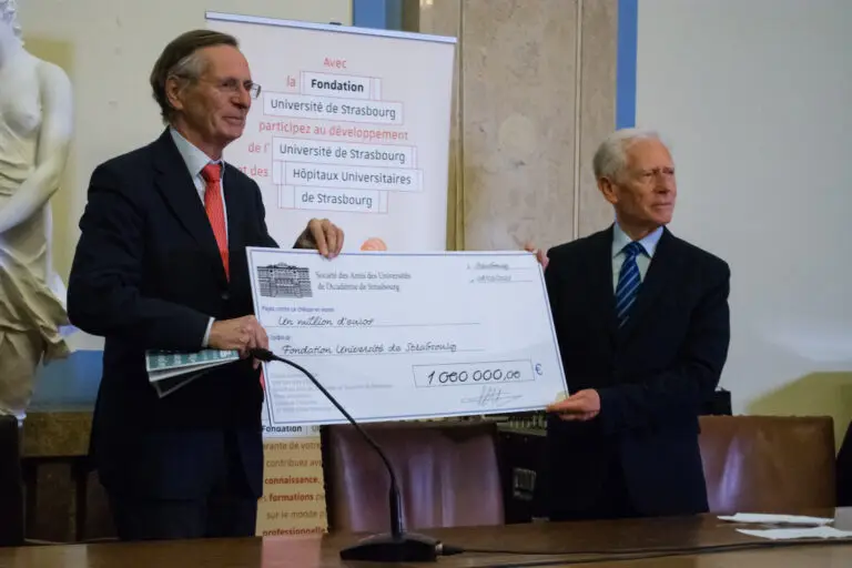 Deux hommes en costume brandissent un chèque géant d'un million d'euros, symbolisant le généreux don d'un million d'euros de la Société des Amis des Universités et de l'Académie de Strasbourg, remis à la Fondation de l'Université de Strasbourg lors d'un événement organisé en salle.