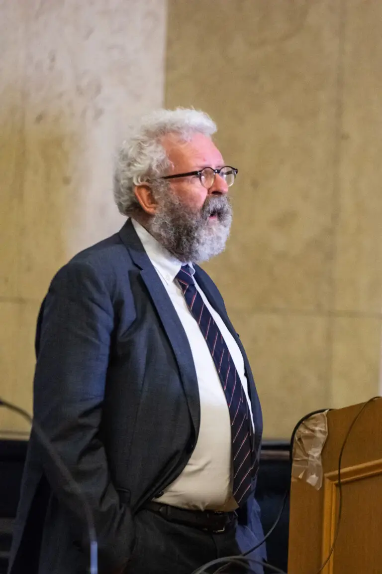 Un homme d'un certain âge, en costume-cravate, se tient debout derrière un pupitre et prend la parole, avec un arrière-plan neutre derrière lui — peut-être aborde-t-il des sujets liés à l'Université de Strasbourg ou parle-t-il de mécénat, à l'instar de ce que ferait l'historien Georges Bischoff.