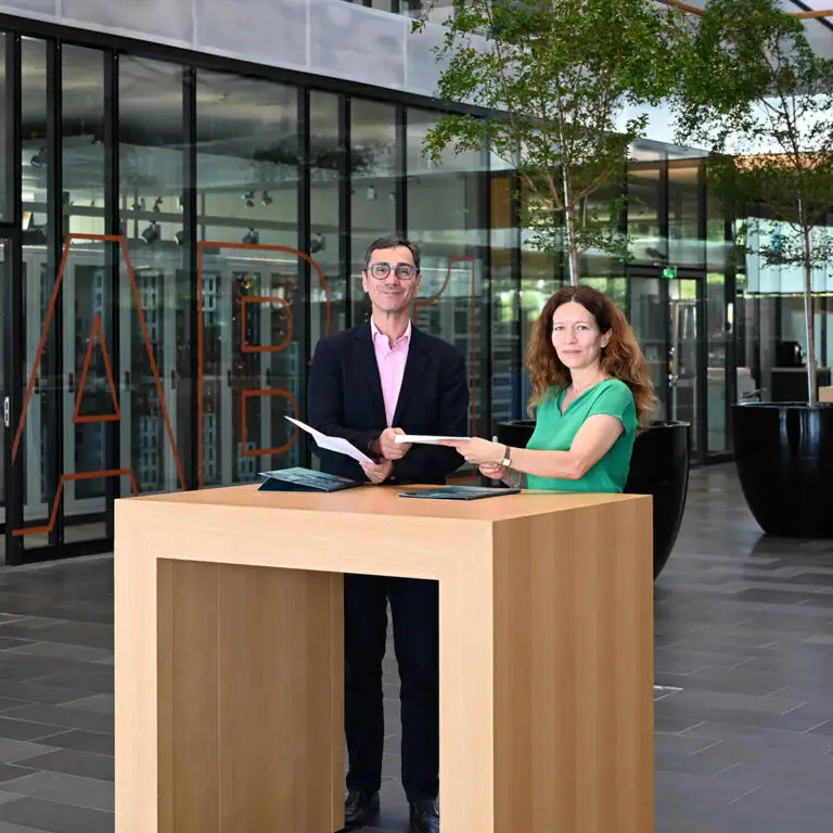 Deux personnes se tiennent debout devant une table en bois, dans un bâtiment moderne aux parois vitrées, documents à la main et face à la caméra, soulignant ainsi la collaboration entre Hager Group et l'Université de Strasbourg. Ce moment incarne l'esprit d'une entreprise familiale engagée dans des partenariats universitaires porteurs de sens.