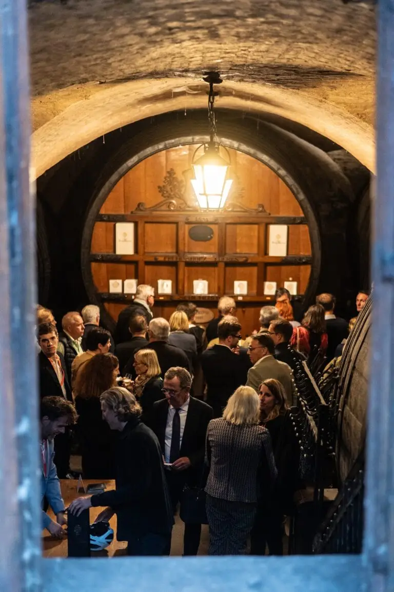 Un groupe de personnes se rassemble dans une cave à vin faiblement éclairée, avec de grands fûts et des plafonds voûtés, et discute sous une suspension lors d'un événement spécial organisé par les Hôpitaux Universitaires de Strasbourg en l'honneur de leurs précieux donateurs.