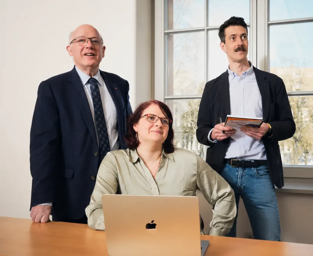 Trois personnes en tenue professionnelle se trouvent dans un bureau ; l'une d'elles est assise à une table devant un ordinateur portable, tandis que les deux autres se tiennent debout à proximité et discutent de ce qui semble être un projet pour la fondation de leur entreprise. À l'arrière-plan, on aperçoit une grande fenêtre donnant sur des arbres et un immeuble.