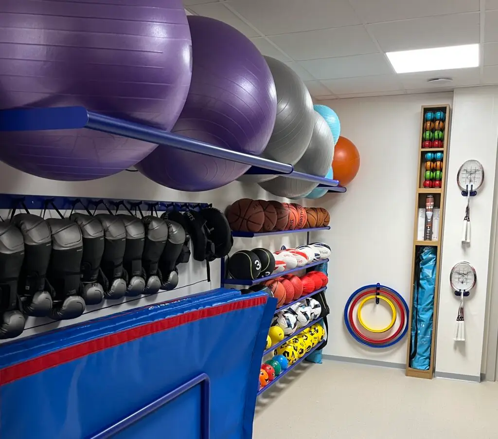 Un local de rangement de la salle de sport contenant des ballons médicinaux, des ballons de basket, des gants de boxe, des raquettes, des cerceaux, des frites en mousse et divers équipements sportifs — dont beaucoup ont été reçus grâce à différentes formes de dons —, le tout soigneusement rangé sur des étagères et des supports.
