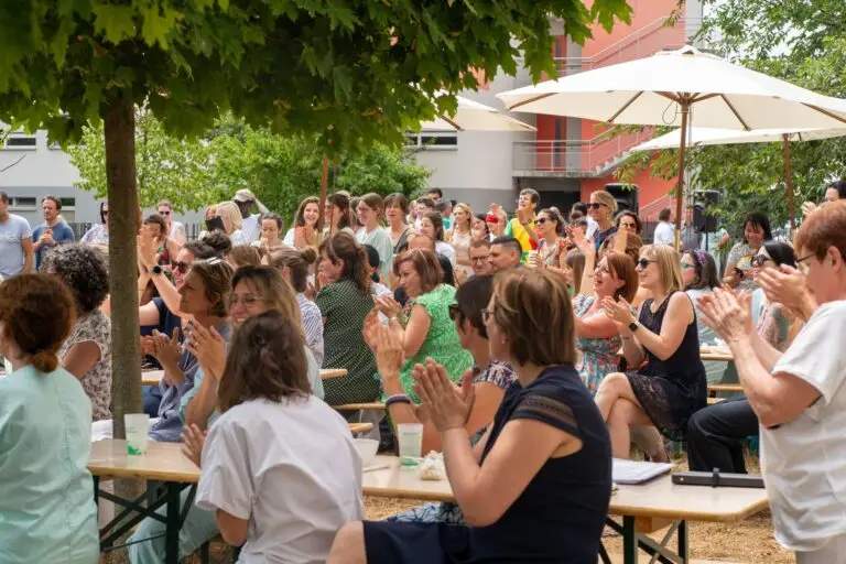 Un groupe de personnes, composé notamment de soignants et d'agents hospitaliers des Hôpitaux universitaires de Strasbourg, est assis ou debout à l'extérieur, à l'ombre des arbres et sous des parasols ; elles applaudissent, toutes tournées dans la même direction, lors d'un événement organisé en journée, avec des bancs et des tables.