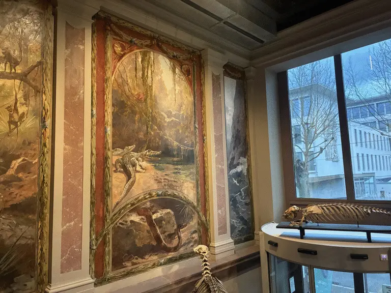 Une fresque murale représentant des dinosaures et des squelettes exposés dans un musée ; de grandes baies vitrées laissent entrevoir des arbres et un bâtiment moderne à l'extérieur, rappelant les expositions immersives du Musée zoologique, où même des créatures telles que les tigres et les pieuvres captivent l'imagination des visiteurs.