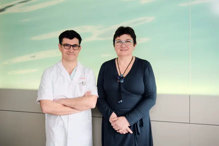 Un homme vêtu d'une blouse blanche se tient aux côtés d'une femme en robe sombre ; tous deux font face à la caméra devant un fond vert pâle, créant une ambiance évoquant la gynécologie et la fertilité.