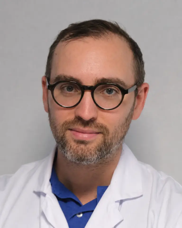 Un homme aux cheveux bruns courts, portant des lunettes et une barbe, revêtu d'une blouse blanche par-dessus une chemise bleue, fait face à la caméra devant un fond clair et uni — incarnant un professionnel voué à la protection des patients.