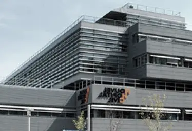 Bâtiment moderne de couleur grise, aux lignes horizontales, doté de nombreuses fenêtres et d'une enseigne orange et noire indiquant « La Fondation » près de l'entrée.