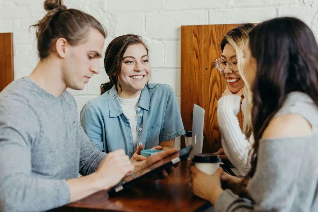 Quatre personnes sont assises à une table en bois dans un café ; elles discutent et sourient tout en utilisant une tablette et en tenant des tasses de café.