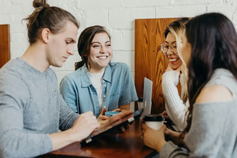 Quatre personnes sont assises à une table en bois dans un café ; elles discutent et sourient tout en utilisant une tablette et en tenant des tasses de café.