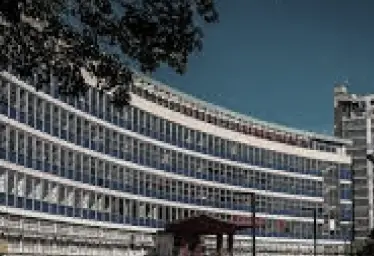 Un grand bâtiment moderne aux lignes courbes, doté de nombreuses baies vitrées et de panneaux bleus et blancs, vu de l'extérieur par temps clair. On aperçoit au premier plan des branches d'arbres et une petite construction.