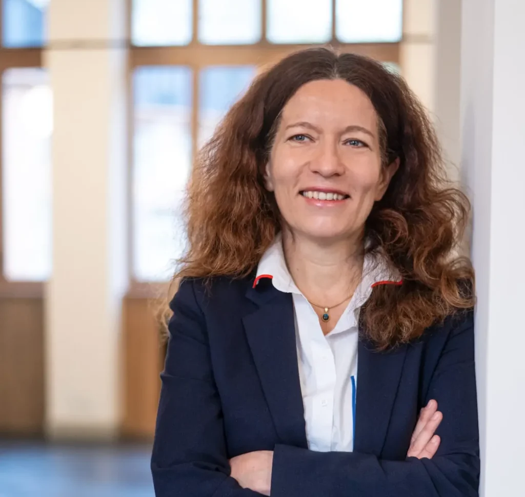 Une femme aux cheveux bruns bouclés, vêtue d'un blazer bleu marine par-dessus une chemise blanche, se tient debout à l'intérieur, les bras croisés, et sourit à la caméra, rayonnant de l'assurance de quelqu'un qui se consacre pleinement à la mission de sa fondation.