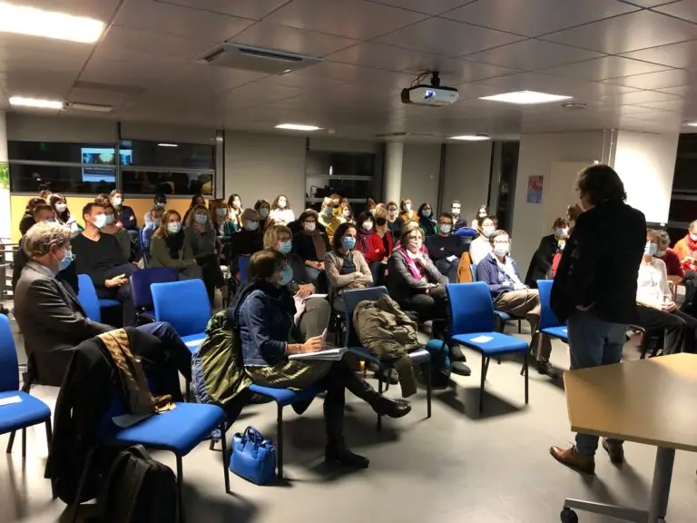 Un groupe de personnes portant des masques est assis sur des chaises bleues, face à un intervenant, dans une salle de classe ou de réunion éclairée par des néons, et assiste à l’un des cycles de conférences organisés par les Hôpitaux universitaires de Strasbourg.