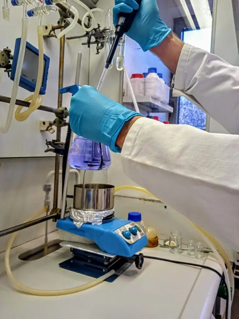 Des mains gantées, vêtues d'une blouse de laboratoire, utilisent une pipette pour verser un liquide dans un bécher contenant une solution violette placée sur un agitateur magnétique, au milieu d'équipements de laboratoire — illustrant ainsi les techniques innovantes en matière de bio-recyclage des plastiques soutenues par les fonds Soprema.