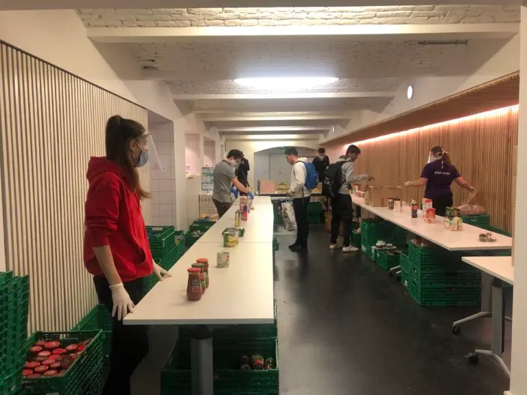 Des personnes portant des masques et des gants trient des conserves sur des tables, dans une pièce bien éclairée où se trouvent des caisses vertes et un plafond en briques blanches, afin de venir en aide aux étudiants de l’Université de Strasbourg grâce aux fonds d’urgence.
