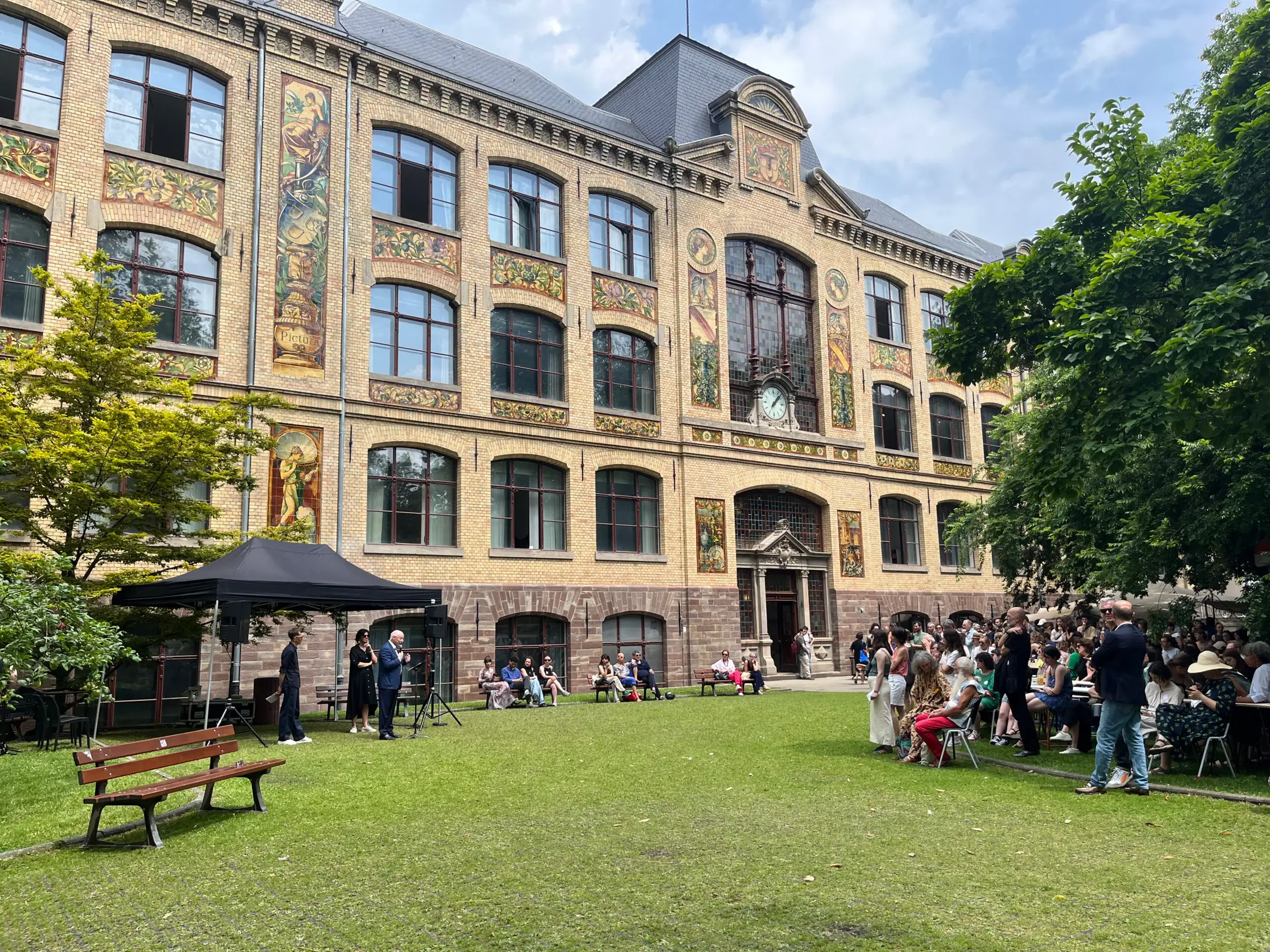 Les gens se rassemblent sur la pelouse devant « La Fondation », un grand bâtiment historique aux fenêtres cintrées et à la façade ornée de fresques.