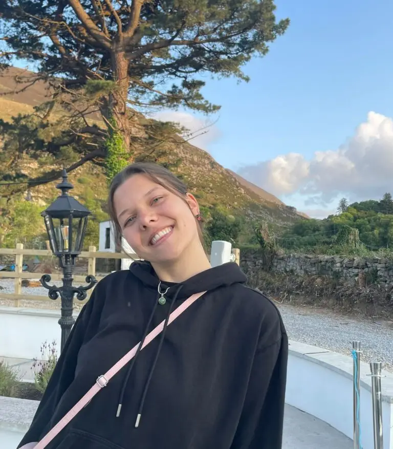 Une personne vêtue d’un sweat à capuche noir se tient à l’extérieur, souriante, avec en arrière-plan une montagne, un arbre et un lampadaire sous un ciel bleu : un cadre idéal pour partager un témoignage sur l’impact qu’a eu l’obtention d’une bourse offerte par de généreux donateurs.