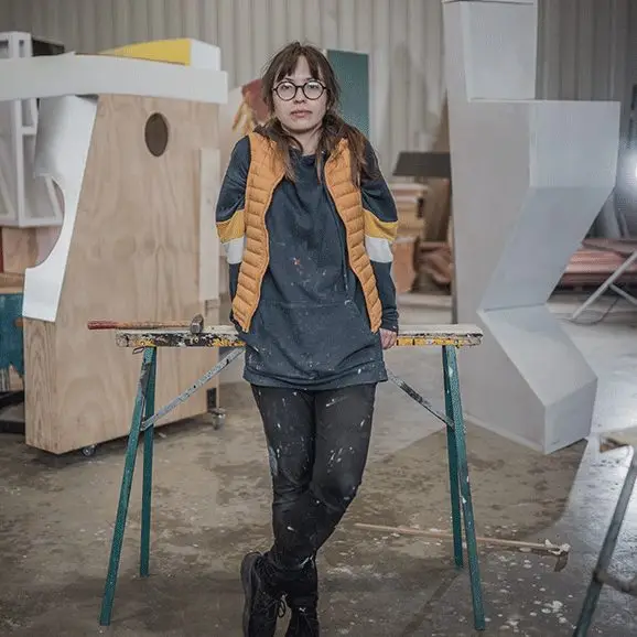 Une personne vêtue d'habits éclaboussés de peinture et portant des lunettes est adossée à un établi dans un atelier d'art, entourée de grandes sculptures abstraites et de structures en bois, incarnant l'esprit des jeunes artistes soutenus par des initiatives telles que la bourse XXLumideco.