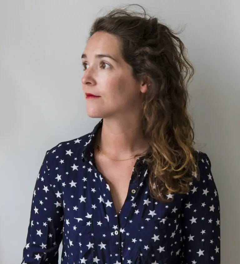 Une femme aux longs cheveux ondulés, vêtue d'un t-shirt bleu marine orné d'étoiles blanches, regardant vers la gauche sur un fond clair et uni — un portrait évocateur qui rappelle le style de Louise Desrosiers à la Haute École des Arts du Rhin.