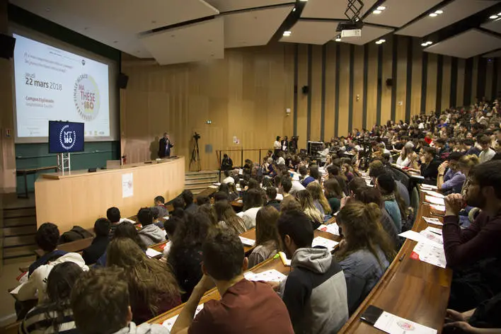 Un grand amphithéâtre rempli d'étudiants assis et écoutant une présentation au Jardin des Sciences, avec Françoise Paya et une autre personne debout à un pupitre près d'un écran de projection, mettant en avant les contributions d'un mécène.